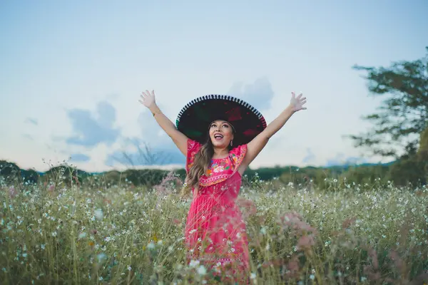 Mexican woman wearing typical Mexican hat and dress, raising her arms with joy. Portrait in a country scene.