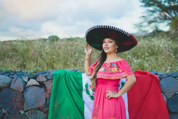 Mexican woman wearing traditional hat and dress next to Mexican flag. Outdoor portrait next to stone wall.