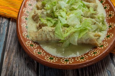 Green Enchiladas with lettuce served in a clay dish on a wooden table.