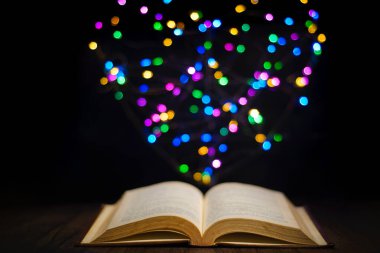 Arka planda bulanık ışıklar olan ahşap masa üzerinde eski açık bir kitap kalp şekli oluşturuyor. Seçici odak.