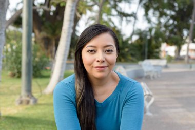 Gülümseyen ve kameraya bakan genç bir kadın, arka planda yeşil çimenler ve ağaçlarla kaplı bir parkta.