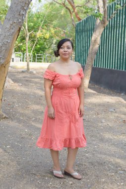 Koyu renk saçlı genç bir kadın mercan rengi bir elbise giyerek gülümsüyor..