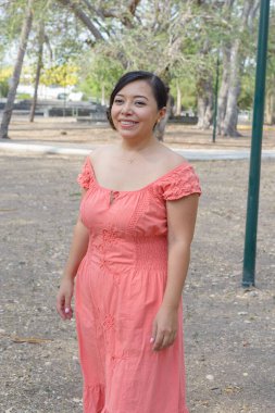 Koyu renk saçlı genç bir kadın mercan rengi bir elbise giyerek gülümsüyor..