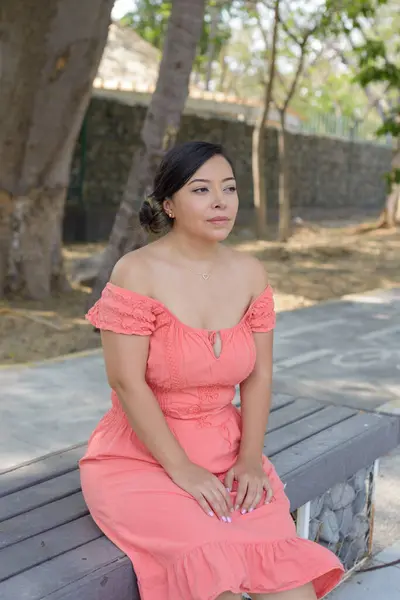 Mercan elbiseli bir kadın düşünceli bir ifadeyle bankta oturuyor.