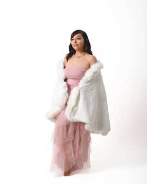 Kendine güvenen bir kadın, sade, açık gri bir arka fona karşı duruyor. Lüks, beyaz, sahte kürk bir pançoyla süslenmiş, narin pembe bir eteği var..