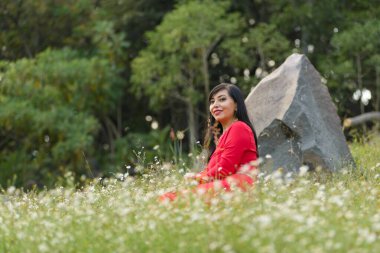 Canlı kırmızı bir elbise giyen neşeli bir kadın kır çiçeklerinin arasında oturuyor..