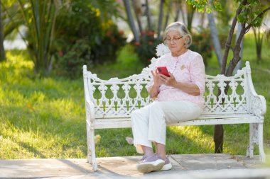 Yaşlı, gri saçlı, gözlüklü bir kadın bir bankta oturuyor, akıllı telefonuna dalmış..