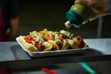 Domates, soğan ve diğer çeşnilerle kaplı sosisli sandviçin üzerine sos döken bir elin yakın çekimi..