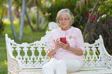 Gri saçlı, gözlüklü, beyaz bir bankta oturan, kırmızı akıllı telefonuna sarılmış son sınıf öğrencisi bir kadın..