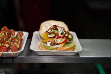Close-up of a freshly prepared burger, garnished with various toppings, resting on a disposable white tray.