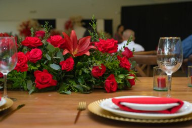 Göz kamaştırıcı bir kırmızı gül süsü ve bir zambak zarif altın şarj aletleri ve kırmızı peçetelerle döşenmiş ahşap bir masayı süsler..