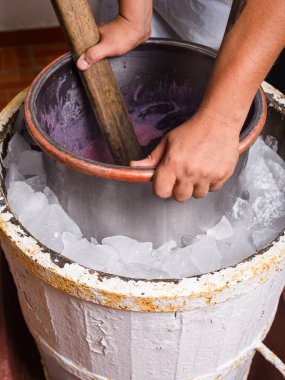 Bir insan, eski moda bir dondurma makinesinin içinde, etrafını buzla çevirmiş, mor bir dondurma yapıyor..