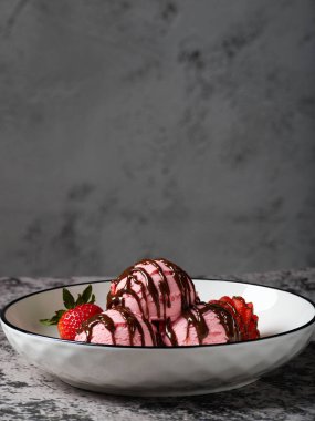 Üç kepçe çilekli dondurma, bol bol çikolata soslu ve beyaz kasede taze çilekle servis ediliyor..