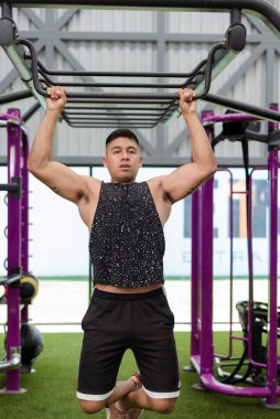 Kaslı kolları olan genç bir adam spor salonunun demir parmaklıklarında barfiks çekiyor. Güç ve zindelik gösterisi yapıyor..