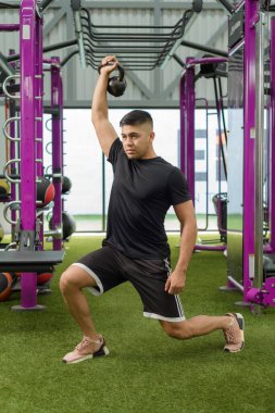 Siyah tişörtlü ve şortlu bir adam modern bir spor salonunda mor spor aletleri ile bir çaydanlık çanı egzersizi yapıyor..