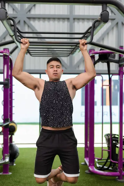 Kaslı kolları olan genç bir adam spor salonunun demir parmaklıklarında barfiks çekiyor. Güç ve zindelik gösterisi yapıyor..