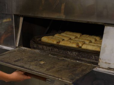 Bir fırıncının elleri dikkatle bir tepsi altın-kahverengiyi, susam tohumunu ticari bir fırına koyar..