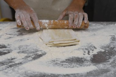 Ustalıkla hamur katmanlarını un kaplı bir yüzeye ahşap bir oklamayla saran eller..