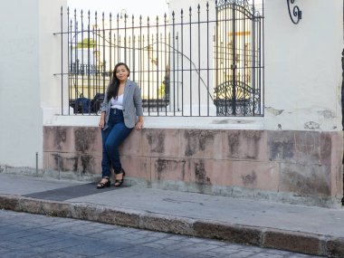 Genç bir kadın tesadüfen alçak bir duvarda oturuyor, yıpranmış taş duvarlar ve arka planda işlenmiş demir çitler..