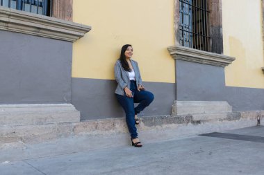 Şık giyinmiş bir kadın ceketini ve kot pantolonunu iki tonlu bir bina duvarına dayamış, rahat ama zarif bir duruşu var..