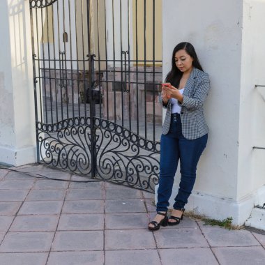 Gingam ceket ve kot giyen genç bir kadın süslü bir demir kapının yanında durup telefonunu kontrol ediyor..