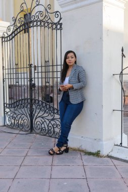 Süslü siyah beyaz spor ceketli ve kot pantolonlu bir kadın süslü demir bir kapının yanında elinde akıllı bir telefon tutuyor..