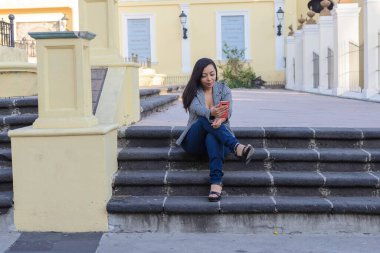 Sarı bir binanın önündeki taş basamaklarda oturan bir kadın dikkatle kırmızı cep telefonuna bakıyor..