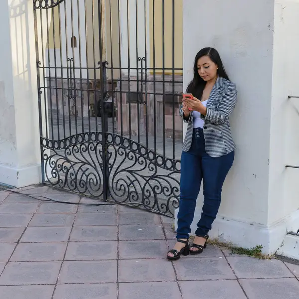 Gingam ceket ve kot giyen genç bir kadın süslü bir demir kapının yanında durup telefonunu kontrol ediyor..