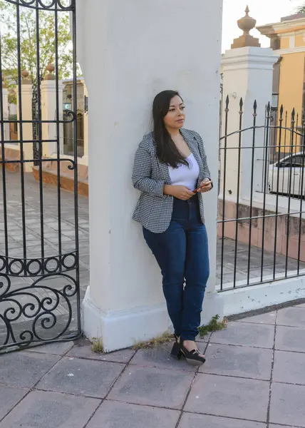 Süslü siyah beyaz spor ceketli ve kot pantolonlu bir kadın süslü demir bir kapının yanında elinde akıllı bir telefon tutuyor..