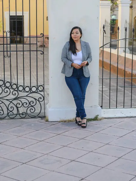 Süslü siyah beyaz spor ceketli ve kot pantolonlu bir kadın süslü demir bir kapının yanında elinde akıllı bir telefon tutuyor..