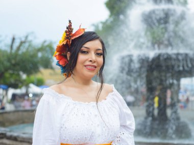 Genç bir kadın sıcak bir şekilde gülümsüyor, beyaz bir göz dirseği ve canlı bir sonbahar çiçeği başlığıyla süslenmiş. Arkaplanda bulanık bir çeşme var.