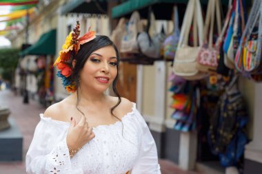Güzel, beyaz işlemeli bir elbise ve çiçekli bir taç giyen genç bir kadın canlı bir Meksika pazarının yanında renkli çantalar ve pançolar sergiliyor..