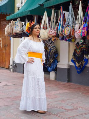 Güzel, beyaz işlemeli bir elbise ve çiçekli bir taç giyen genç bir kadın canlı bir Meksika pazarının yanında renkli çantalar ve pançolar sergiliyor..