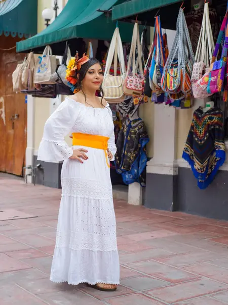 Güzel, beyaz işlemeli bir elbise ve çiçekli bir taç giyen genç bir kadın canlı bir Meksika pazarının yanında renkli çantalar ve pançolar sergiliyor..