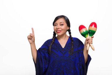 Gülümseyen bir kadın, desenli mavi bir panço ile süslenmiş, marakas tutuyor ve yukarı doğru işaret ediyor..