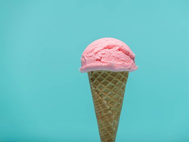 Bir kepçe pembe dondurma gevrek bir waffle külahının üzerinde, canlı bir turkuaz arka planda duruyor..