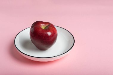 Canlı kırmızı bir elma, siyah kenarlı, küçük beyaz bir tabakta zarif bir şekilde durur..