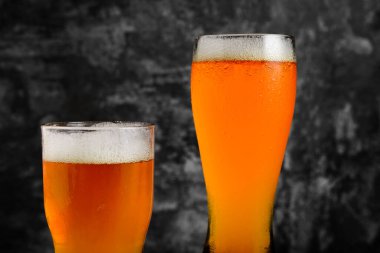 İki bardak kehribar birası, biri uzun, biri kısa koyu renkli bir arka planda duruyor..