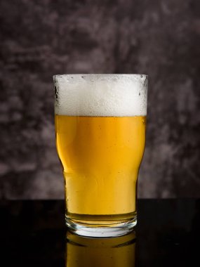 Kalın bir köpük baş ile kaplı, ferahlatıcı bir altın bira, koyu, yansıtıcı bir yüzey üzerinde oturur bulanık bir arka plan karşı.