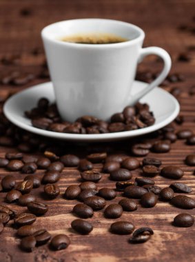 Koyu kahve ve espresso fincanı, ahşap bir yüzeyde kavrulmuş fasulyelerle çevrili bir çay tabağının üzerinde oturur. Seçici odak.