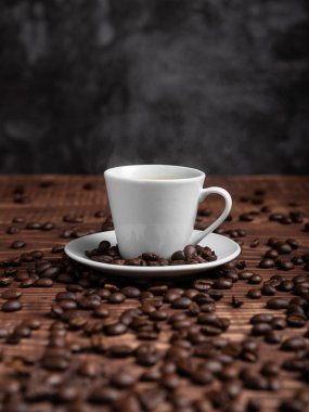 Koyu kahve ve espresso fincanı, ahşap bir yüzeyde kavrulmuş fasulyelerle çevrili bir çay tabağının üzerinde oturur. Seçici odak.