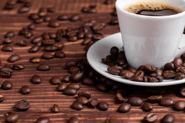 Koyu kahve ve espresso fincanı, ahşap bir yüzeyde kavrulmuş fasulyelerle çevrili bir çay tabağının üzerinde oturur. Seçici odak.