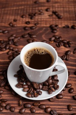 Kahverengi ahşap bir yüzey üzerinde kahve çekirdekleri ile çevrili, beyaz bir fincan üzerinde koyu kavrulmuş bir kahve.