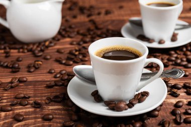 Kahverengi ahşap bir yüzey üzerinde kahve çekirdekleri ile çevrili, beyaz bir fincan üzerinde koyu kavrulmuş bir kahve.