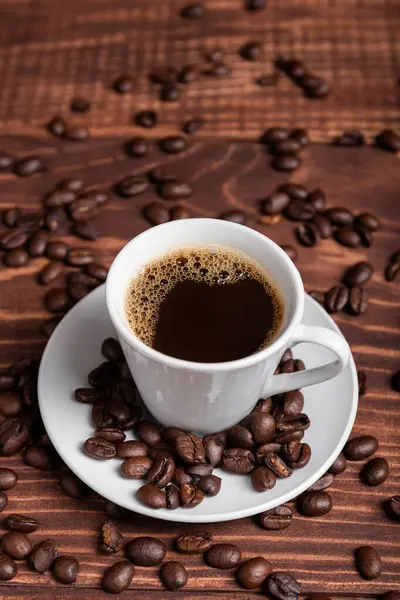 Kahverengi ahşap bir yüzey üzerinde kahve çekirdekleri ile çevrili, beyaz bir fincan üzerinde koyu kavrulmuş bir kahve.