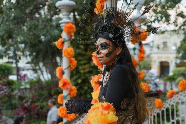 Kafatası makyajı ve çarpıcı bir başlıkla süslenmiş bir kadın canlı turuncu kadife çiçekleri arasında duruyor.