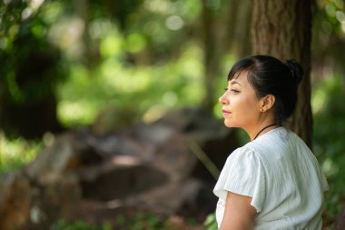 Bir kadın yemyeşil bir ormanda huzur içinde oturuyor, bakışları mesafeye bakıyor. Güneş ışığı yaprakların arasından süzülür.