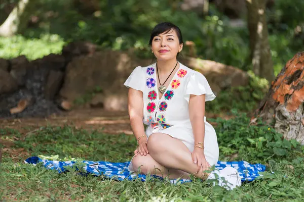 Beyaz işlemeli elbiseli bir kadın, yeşil bir parkta, mavi ve beyaz bir battaniyenin üzerinde huzur içinde oturur ve sessizce yukarı bakar..