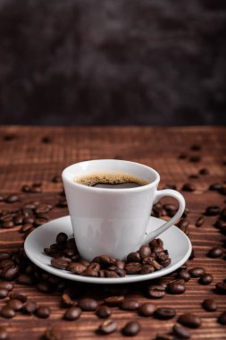 Kahverengi ahşap bir yüzey üzerinde kahve çekirdekleri ile çevrili, beyaz bir fincan üzerinde koyu kavrulmuş bir kahve.