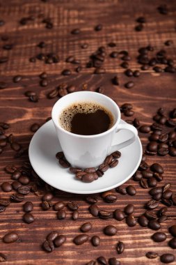 Kahverengi ahşap bir yüzey üzerinde kahve çekirdekleri ile çevrili, beyaz bir fincan üzerinde koyu kavrulmuş bir kahve.